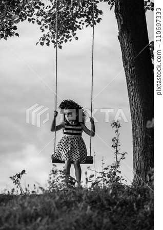 A small and beautiful girl in a dress is swinging on a swing. A small and beautiful girl in a dress is swinging on a swing. 110697333