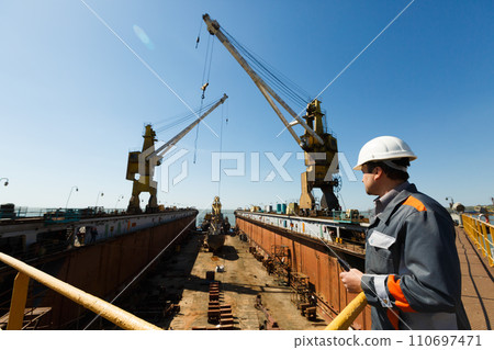 Professional overseeing extensive shipbuilding process, massive cranes erect vessel in dry dock against a clear sky. Maritime engineer in hardhat and reflective vest monitors construction. Professional overseeing extensive shipbuilding process, massive cranes erect vessel in dry dock against a clear sky. Maritime engineer in hardhat and reflective vest monitors construction. 110697471