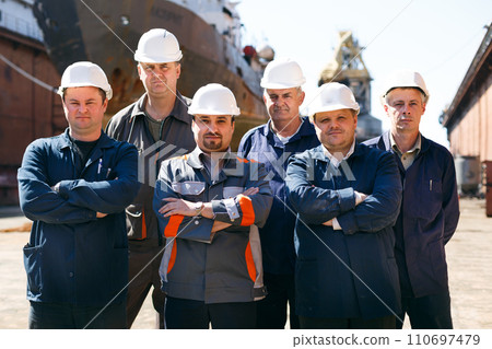 Shipyard workers at float dock wearing hard hats, stand arms-crossed. Male repair team ready for maintenance work on vessel. Maritime industry, professional ship fix group, outdoor labor day scene. 110697479