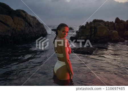 Attractive female model enjoys nighttime dip in sea illuminated by crimson lights, her style radiant in designer swimwear. The tranquil waters reflect her poised elegance amidst rocky seaside. 110697485