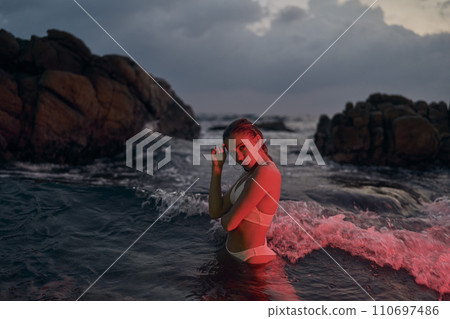 Attractive woman enjoys night swimming in ocean illuminated by red light. Fashionable female in swimwear experiences nocturnal water adventure. Elegant lady in glowing sea, beauty concept in nature. 110697486