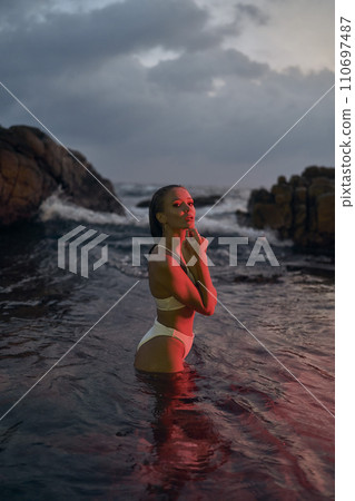 Attractive female enjoys nocturnal swim in sea under red illumination. Young woman in bikini experiences tranquil night dip amidst marine ambiance. Oceanic holiday, leisure travel for relax. 110697487