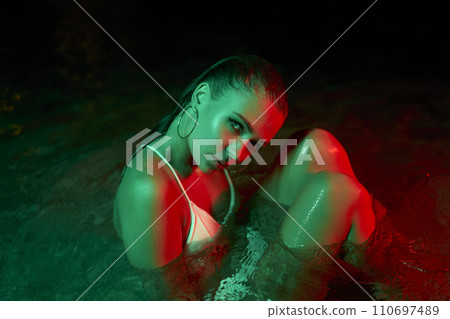 Woman sits in a water lagoon at night, enigmatic under green and red hues. Mysterious atmosphere, fusion of nature and futuristic aesthetic. Feminity, beauty on a night swim session. Swimwear fashion. Woman sits in a water lagoon at night, enigmatic under green and red hues. Mysterious atmosphere, fusion of nature and futuristic aesthetic. Feminity, beauty on a night swim session. Swimwear fashion. 110697489
