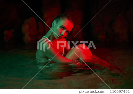 Woman sits in water at night, illuminated by green red light. Female model in aquatic environment, creative lighting, night photography. Concept beauty, self, wet woman, lagoon scene. Woman sits in water at night, illuminated by green red light. Female model in aquatic environment, creative lighting, night photography. Concept beauty, self, wet woman, lagoon scene. 110697491