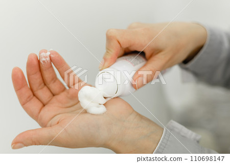 Hands of a pedicure master in a bathrobe and black gloves squeeze cream from the bottle. 110698147