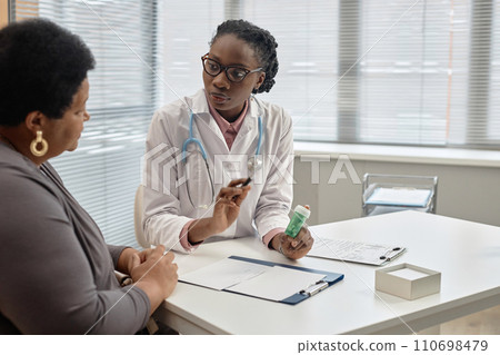 Young African American general practitioner explaining medication administration to female patient while sitting at table in hospital Young African American general practitioner explaining medication administration to female patient while sitting at table in hospital 110698479