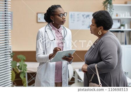 Medium long shot of young African American woman physician in clinic office talking to senior female patient and holding clipboard Medium long shot of young African American woman physician in clinic office talking to senior female patient and holding clipboard 110698513