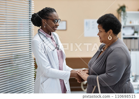 Side view of cheerful Black woman doctor greeting mature female patient while shaking hands in clinic 110698515