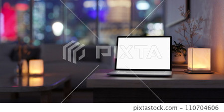 A white-screen laptop computer and a table lamp on a table in a modern, dark living room at night. A white-screen laptop computer and a table lamp on a table in a modern, dark living room at night. 110704606