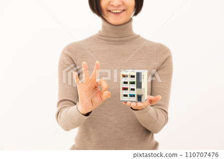 A middle-aged woman holding a model of an apartment building and making a circle sign A middle-aged woman holding a model of an apartment building and making a circle sign 110707476