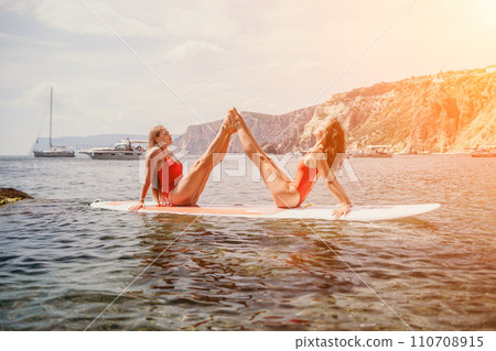 Woman sup yoga. Two happy sporty women practising yoga pilates on paddle sup surfboard. Female stretching doing workout on sea water. Modern individual female hipster outdoor summer sport activity. 110708915