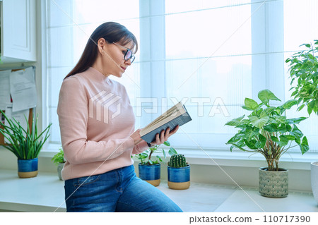 Middle-aged woman reading fiction book at home, at home near window 110717390