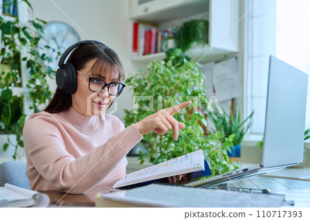Woman teacher in headphones with textbook working online using computer Woman teacher in headphones with textbook working online using computer 110717393