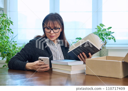 Woman with smartphone unpacking box with books, online shopping 110717409