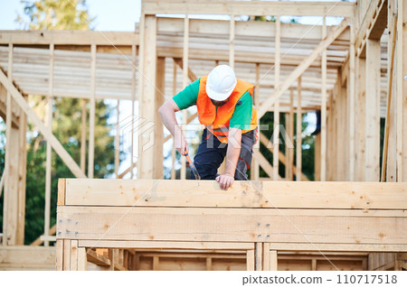 Carpenter constructing two-story wooden frame house. Bearded man hammering nails into structure while wearing protective helmet and construction vest. Concept of modern ecological construction. Carpenter constructing two-story wooden frame house. Bearded man hammering nails into structure while wearing protective helmet and construction vest. Concept of modern ecological construction. 110717518