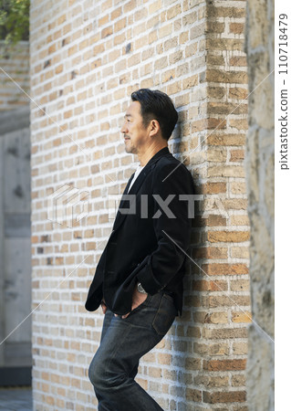 A man in his mid-50s wearing a black jacket leaning against a brick wall. 110718479