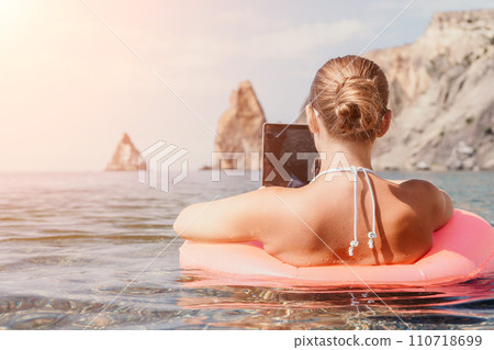 Woman freelancer works on laptop swimming in sea on pink inflatable ring. Happy tourist in sunglasses floating on inflatable donut and working on laptop computer in calm ocean. Remote working anywhere 110718699