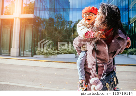 Cute laughing daughter on a piggy back ride with her brunette happy mother in autumn time with modern building at city street on background. 110719114
