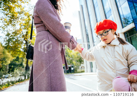 Cute little girl in red wear beret and coat in eyeglasses, whirling frolicking and playing with her unrecognizable mother at autumn sunny day city street outdoor. 110719116