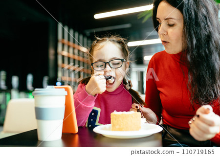 Happy family moments concept. Cute little daughter in eyeglasses and her mother eating cake at cafe together. 110719165