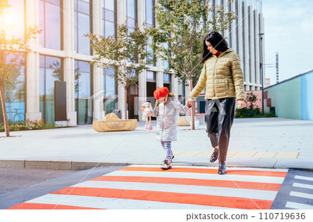 Young caucasian mother crossing street and commuting downtown city with little daughter wearing eyeglasses and red beret in autumn time. Full height. Young caucasian mother crossing street and commuting downtown city with little daughter wearing eyeglasses and red beret in autumn time. Full height. 110719636