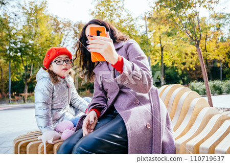 Brunette young mother showing cartoons to disability little adorable girl on smartphone. Woman recording family video with small preschool daughter on mobile phone sitting on bench at park outdoor. Brunette young mother showing cartoons to disability little adorable girl on smartphone. Woman recording family video with small preschool daughter on mobile phone sitting on bench at park outdoor. 110719637