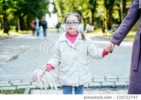 Special need child holding mother's hands and toy wearing headphones having fun in city park in autumn. Special need child holding mother's hands and toy wearing headphones having fun in city park in autumn. 110722747