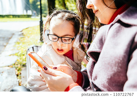 Special need child wearing headphones and happy time to use a smartphone on bench with her mother in sunny park outdoor. 110722767