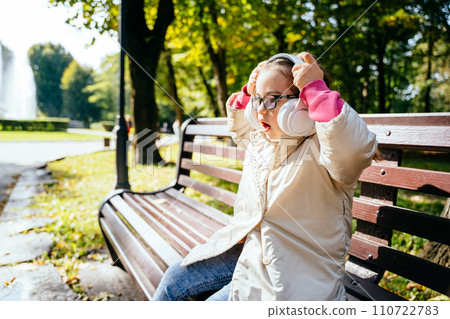 Side view of cute lovely little girl listening music with headphones sitting on the bench at park outdoor. 110722783