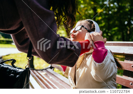 Unrecognizable mother putting headphones her little disabled daughter in eyeglasses for calming outdoor in park. Down Syndrome in daily lives. 110722784