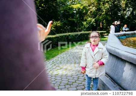 Portrait sad down syndrome girl standing in front of her unrecognizable mother with a sad face. Child with a disability is naughty and does not listen to her mother in park outdoor. Portrait sad down syndrome girl standing in front of her unrecognizable mother with a sad face. Child with a disability is naughty and does not listen to her mother in park outdoor. 110722798