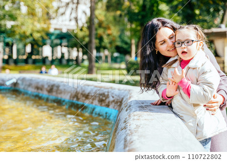 Little girl with special needs enjoy spending time with mother in autumn park outdoor. Child feeling love from parent. Portrait of happy woman with her daughter near fountained. 110722802