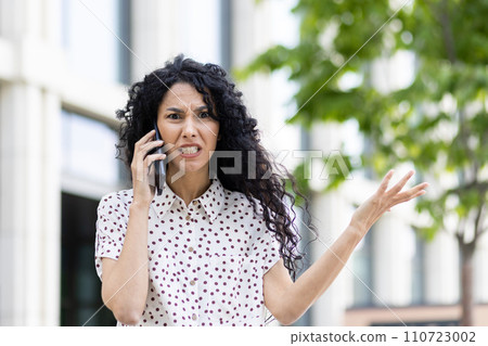 Curly-haired woman in a spotted blouse expressing concern while talking on the mobile phone, an office building backdrop. 110723002