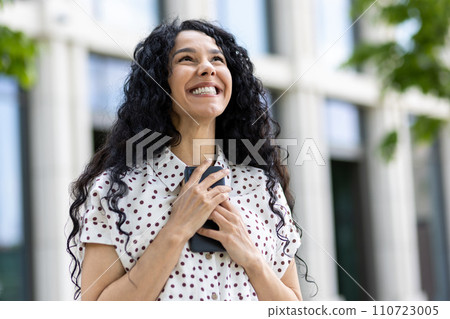 Optimistic young Latin American woman with curly hair smiling outside an office building, reflecting confidence and success. 110723005