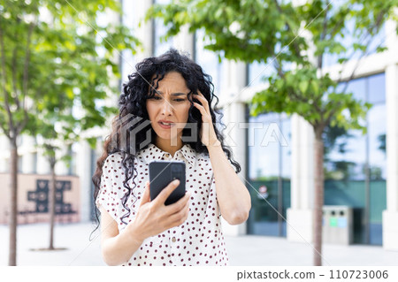 Anxious young woman with curly hair looking at phone screen with a concerned expression, standing outdoors near office buildings. 110723006