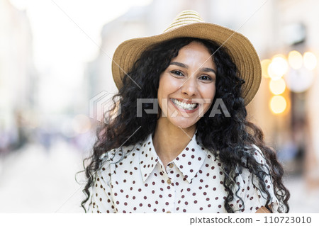 Portrait of a joyful Latina businesswoman with curly hair and a hat, on a city street with a blur background of city lights. 110723010