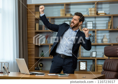 Energetic mature businessman in a smart suit celebrates victory with a fist pump in a well-appointed home office setting. 110723017