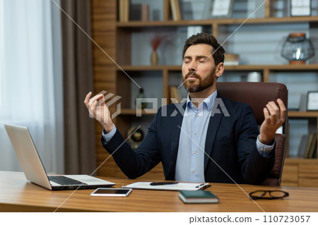 Experienced male professional in a suit practicing mindfulness meditation at his workplace for relaxation. Experienced male professional in a suit practicing mindfulness meditation at his workplace for relaxation. 110723057