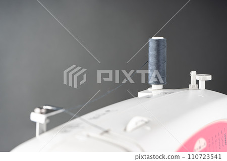 Spool of gray sewing thread stands on top of a white modern sewing machine Spool of gray sewing thread stands on top of a white modern sewing machine 110723541