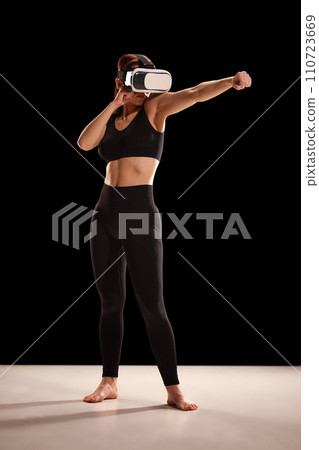 Strong and fit woman in sportswear watching video-training in VR glasses and repeat moves in action against black studio background. 110723669