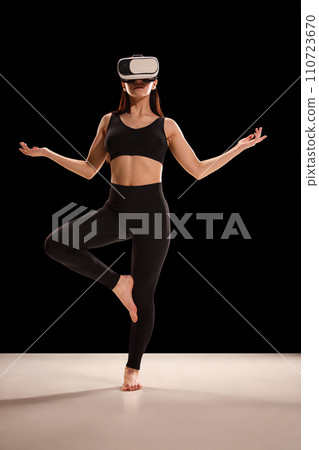 Woman in fitness costume and VR glasses doing yoga exercises against black studio background. Body care and mindfulness. Woman in fitness costume and VR glasses doing yoga exercises against black studio background. Body care and mindfulness. 110723670