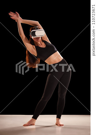 Portrait of young woman in sportwear doing exercises for back in virtual reality glasses against back studio background. Portrait of young woman in sportwear doing exercises for back in virtual reality glasses against back studio background. 110723671