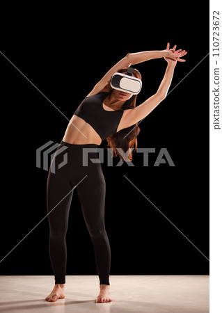 Portrait of young woman in fitness wear doing back exercises in virtual reality glasses against back studio background. 110723672
