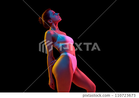 Side view of young woman in white lingerie posing looking up against black studio background in neon light. Side view of young woman in white lingerie posing looking up against black studio background in neon light. 110723673
