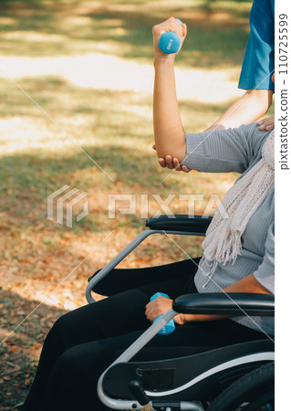 Young nurse or physiotherapist in scrubs helping a happy retired old woman do fitness exercises with light weight dumbbells at home. Concept of physiotherapy for seniors 110725599