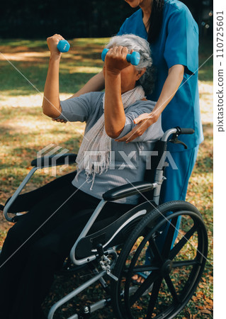 Young nurse or physiotherapist in scrubs helping a happy retired old woman do fitness exercises with light weight dumbbells at home. Concept of physiotherapy for seniors 110725601