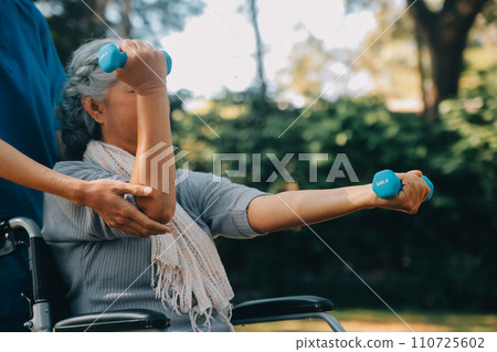 Young nurse or physiotherapist in scrubs helping a happy retired old woman do fitness exercises with light weight dumbbells at home. Concept of physiotherapy for seniors 110725602