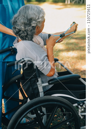 Young nurse or physiotherapist in scrubs helping a happy retired old woman do fitness exercises with light weight dumbbells at home. Concept of physiotherapy for seniors 110725611