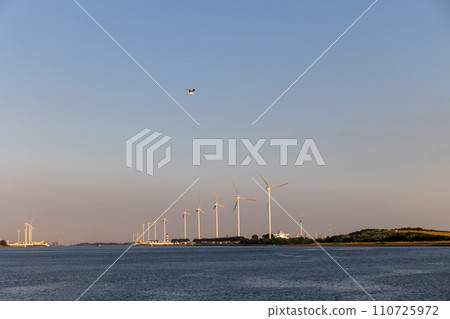 Beautiful windmills in the port of Rotterdam. Beautiful sunset on the sea coast. The Blue North Sea and Water surface. The lighthouse and shore are lit by the sunset sun. 110725972