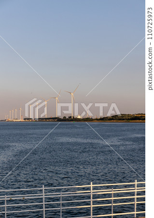 Beautiful windmills in the port of Rotterdam. Beautiful sunset on the sea coast. The Blue North Sea and Water surface. The lighthouse and shore are lit by the sunset sun. Beautiful windmills in the port of Rotterdam. Beautiful sunset on the sea coast. The Blue North Sea and Water surface. The lighthouse and shore are lit by the sunset sun. 110725973